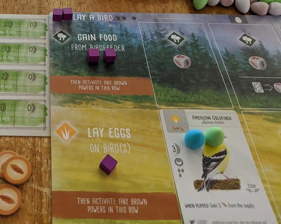 Purple wooden action cubes on Wingspan player mat as well as bird cards and eggs. Goal tracker card and food tokens to the left of the mat.