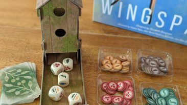 Overhead view of cardboard dice tower that looks like a bird feeder with custom wooden dice in the tray next to food tokens.