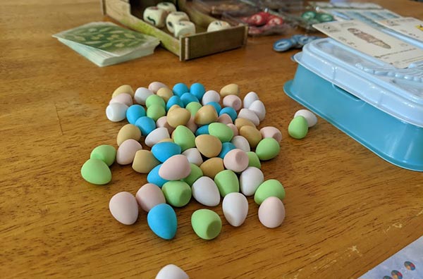 A pile of miniature eggs in white, green, blue, pink, and yellow on table next to other Wingspan game components.